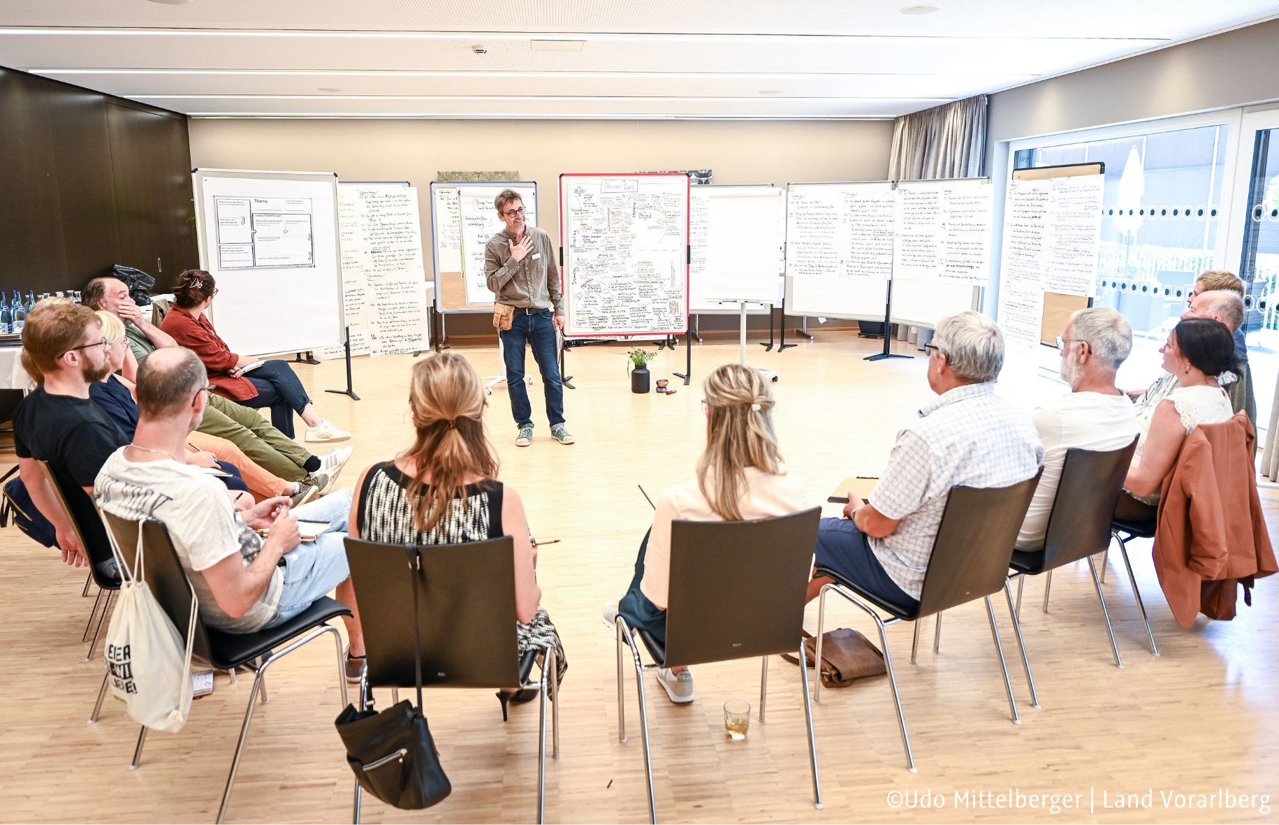 Personen sitzen in einem Stuhlkreis. Beschriebene Flipcharts sind im Hintergrund. Ein Moderator spricht. ©Udo Mittelberger | Land Vorarlberg