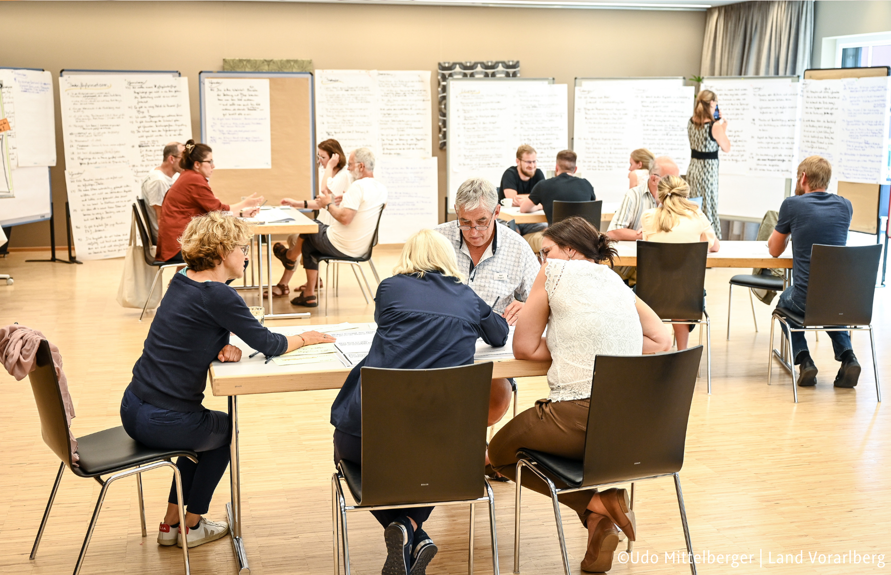 Personen sitzen an Tischen in kleinen Gruppen. Sie sprechen miteinander und schreiben auf Flipcharts. ©Udo Mittelberger | Land Vorarlberg